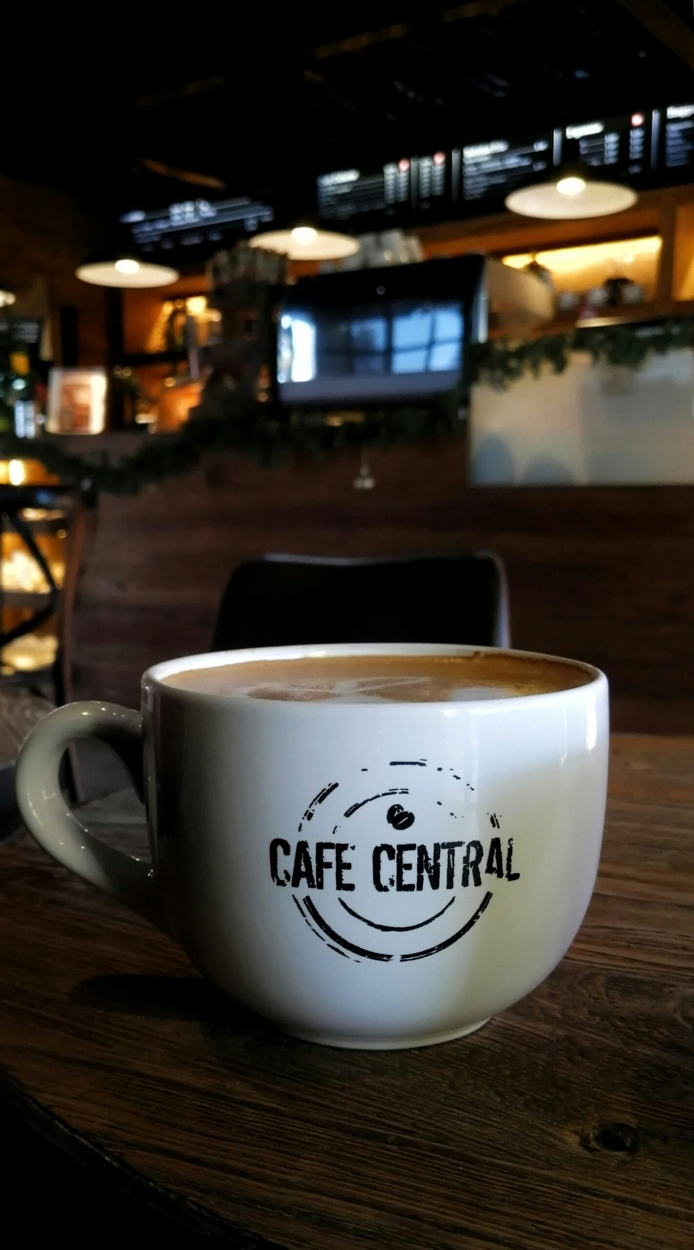 A white mug of latte with the Cafe Central logo on a wooden table in the cafe's dimly lit interior