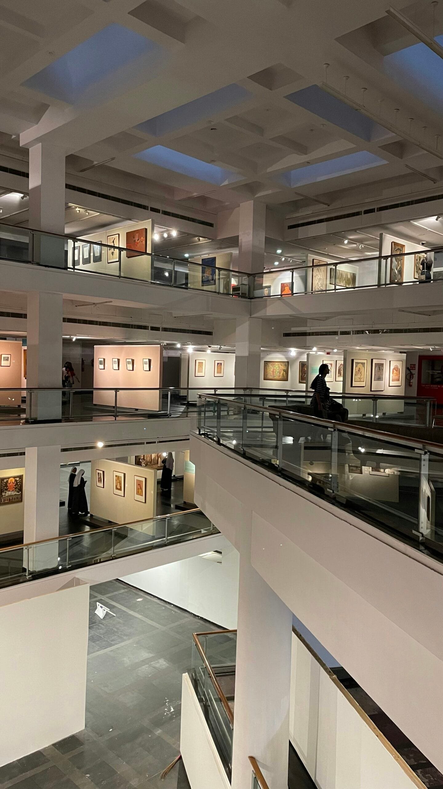 Multi-level interior of a modern art gallery with white pillars, glass railings, and artworks displayed on several floors