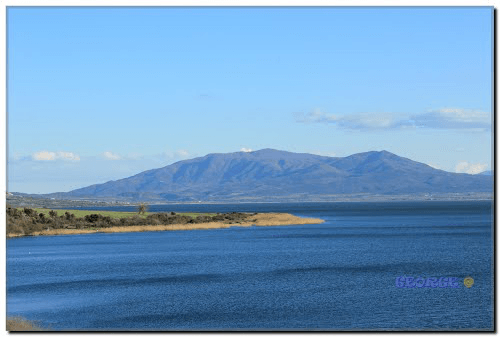 Wide view of Lake Volvi with a reeded shoreline and large mountains in the background under a clear blue sky