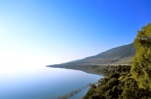 Calm blue waters of Lake Volvi bordered by a green shoreline and a road at the base of a sloping mountain