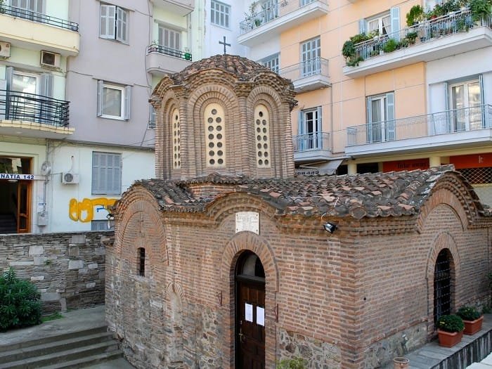 Small Byzantine brick church with a central dome and cross, surrounded by modern apartment buildings
