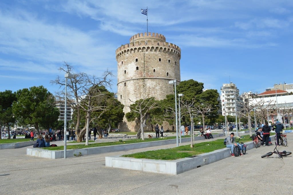 The circular stone White Tower of Thessaloniki with a Greek flag on top, surrounded by a park with people