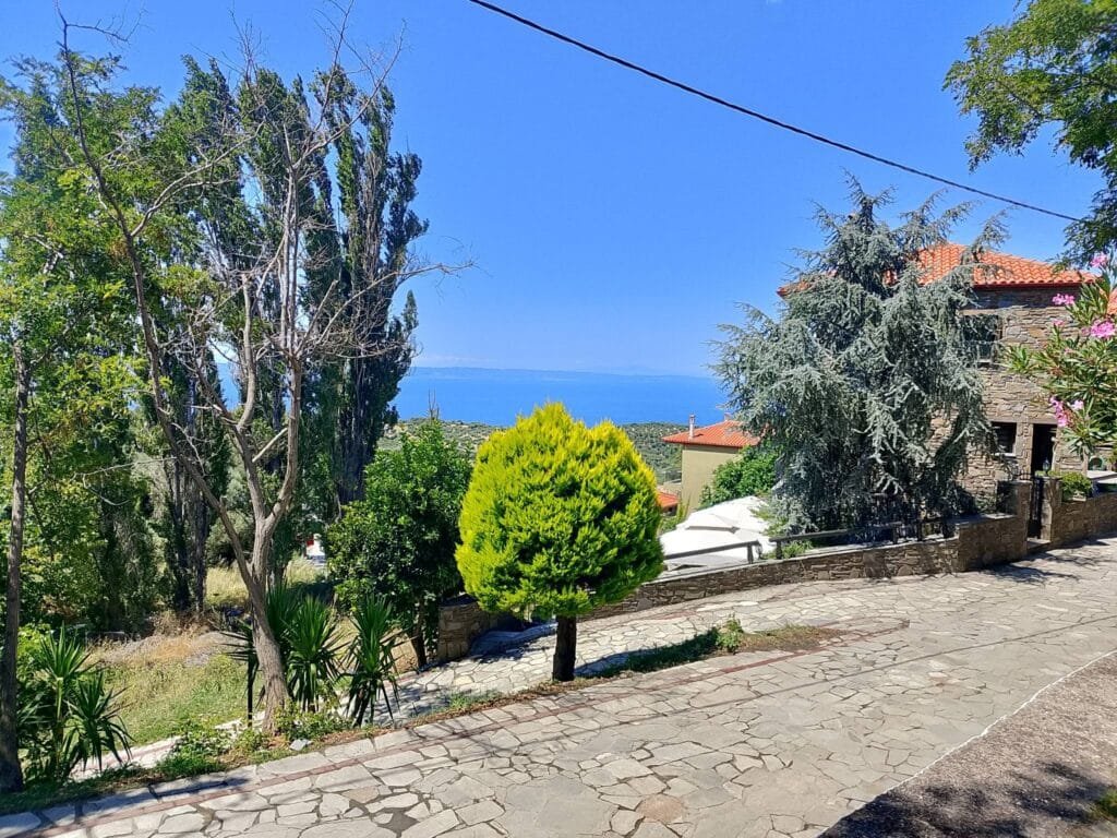 Stone-paved street in Parthenon village with lush trees and a view of the blue Aegean Sea in the distance