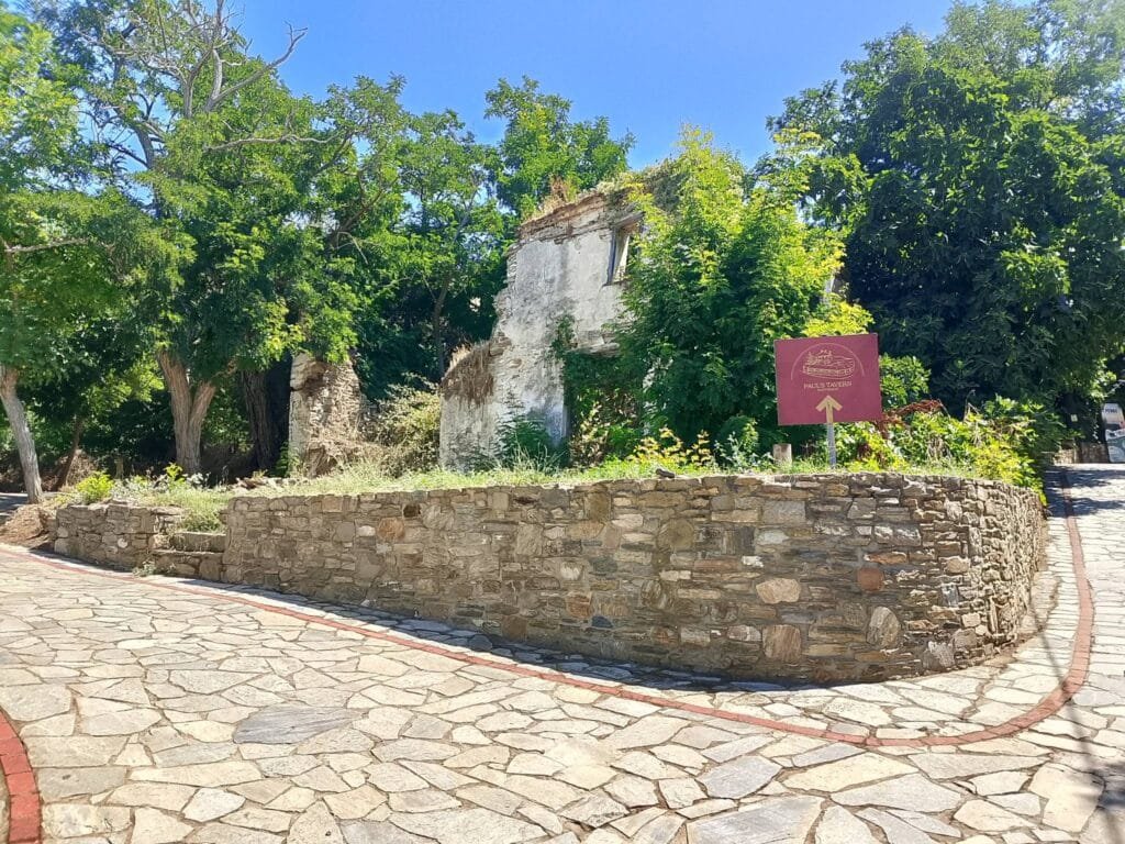 Stone-paved street and low wall in Parthenon village, with a ruined stone building and lush green trees