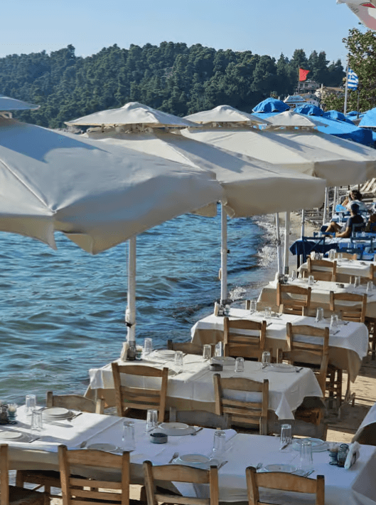 Outdoor restaurant tables with white tablecloths and umbrellas set along the sea shore against forested hills