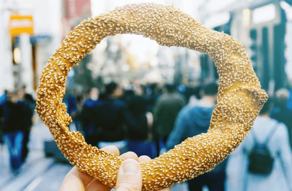 Hand holding a sesame-covered bread ring (koulouri) against a blurred city street background
