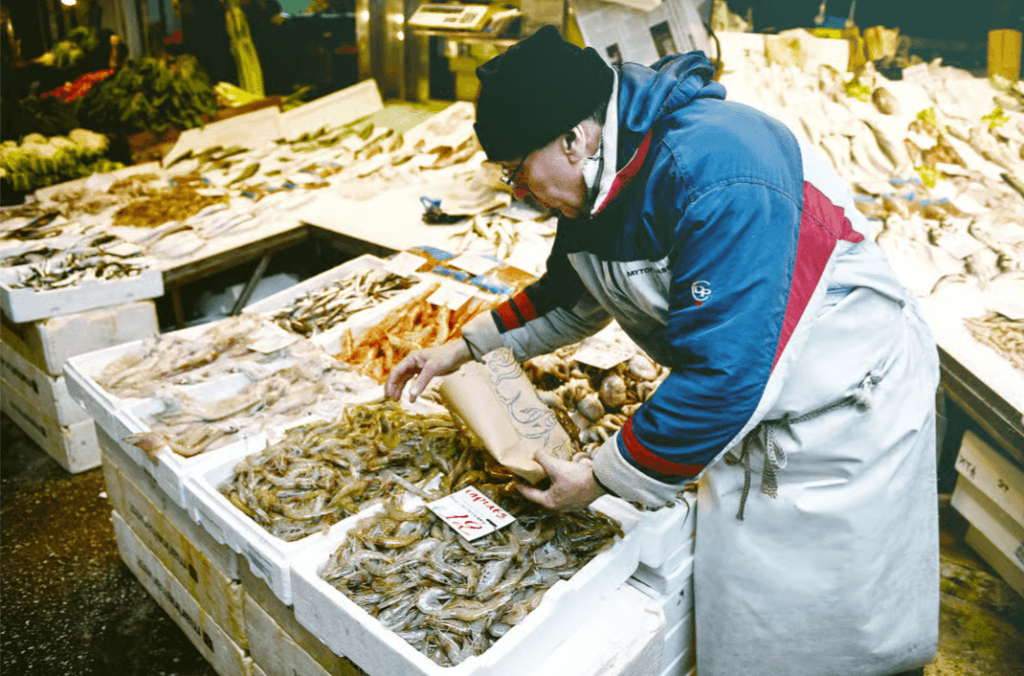 A fishmonger at a market stall selects fresh shrimp from a display of various seafood
