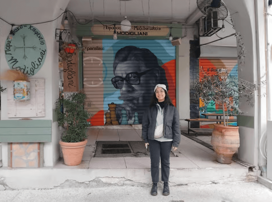 A woman stands in front of a mural of a man with glasses and chess pieces on a shop shutter in Thessaloniki