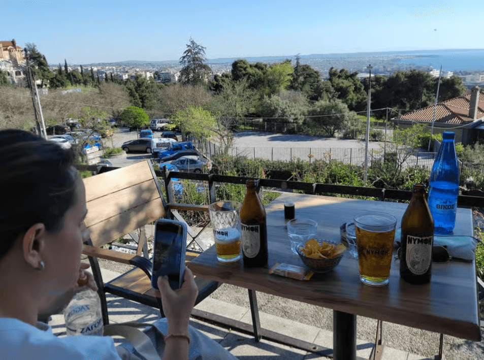 A person at an outdoor cafe table with local beers, overlooking a panoramic view of Thessaloniki and the sea