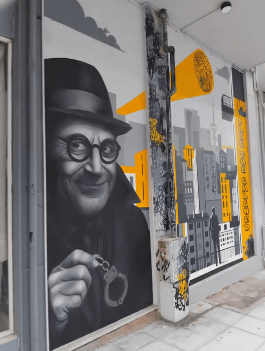 Mural on a city wall featuring a man in a hat and glasses holding handcuffs next to a yellow and grey cityscape