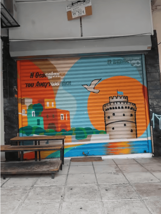 Mural on a shop shutter depicting the White Tower at sunset with the text Η Θεσσαλονίκη του Αναγνωστάκη