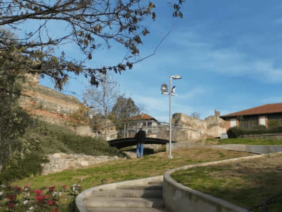 A person crosses a small bridge near the historic Byzantine walls of Thessaloniki under a clear blue sky