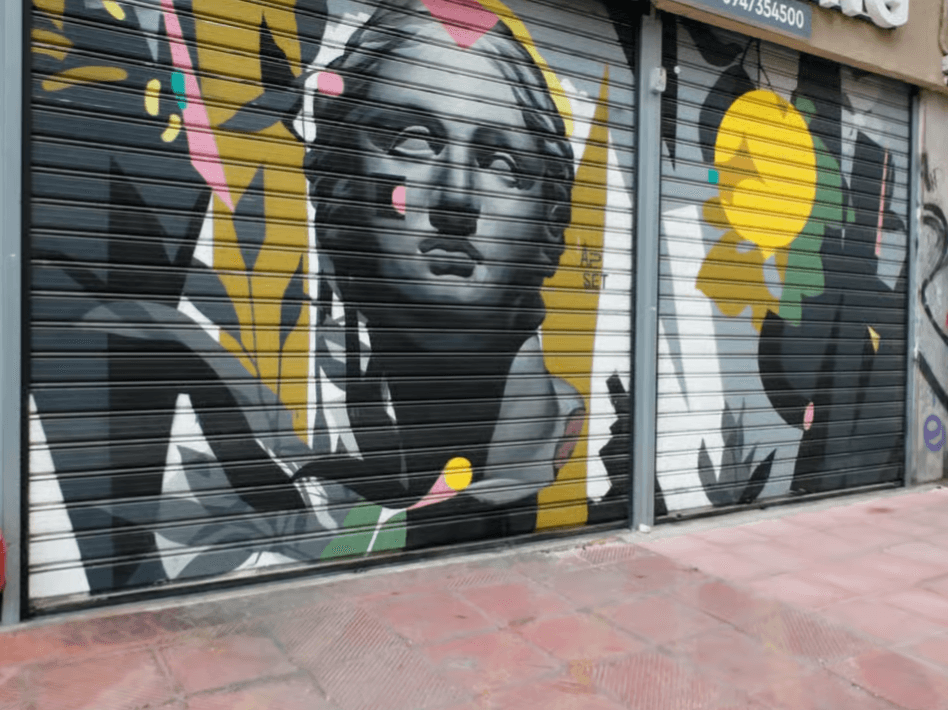 Mural on a metal shop shutter featuring a grayscale classical bust with colorful abstract shapes and patterns