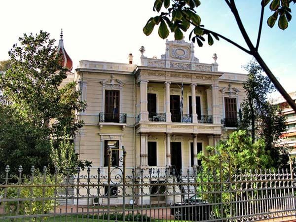 Ornate two-story mansion with classical columns, balconies, and decorative moldings behind an iron fence