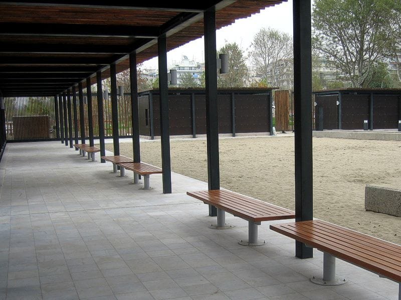 Wooden benches under a long black pergola with a wooden roof, next to a sandy area in the Rose Garden