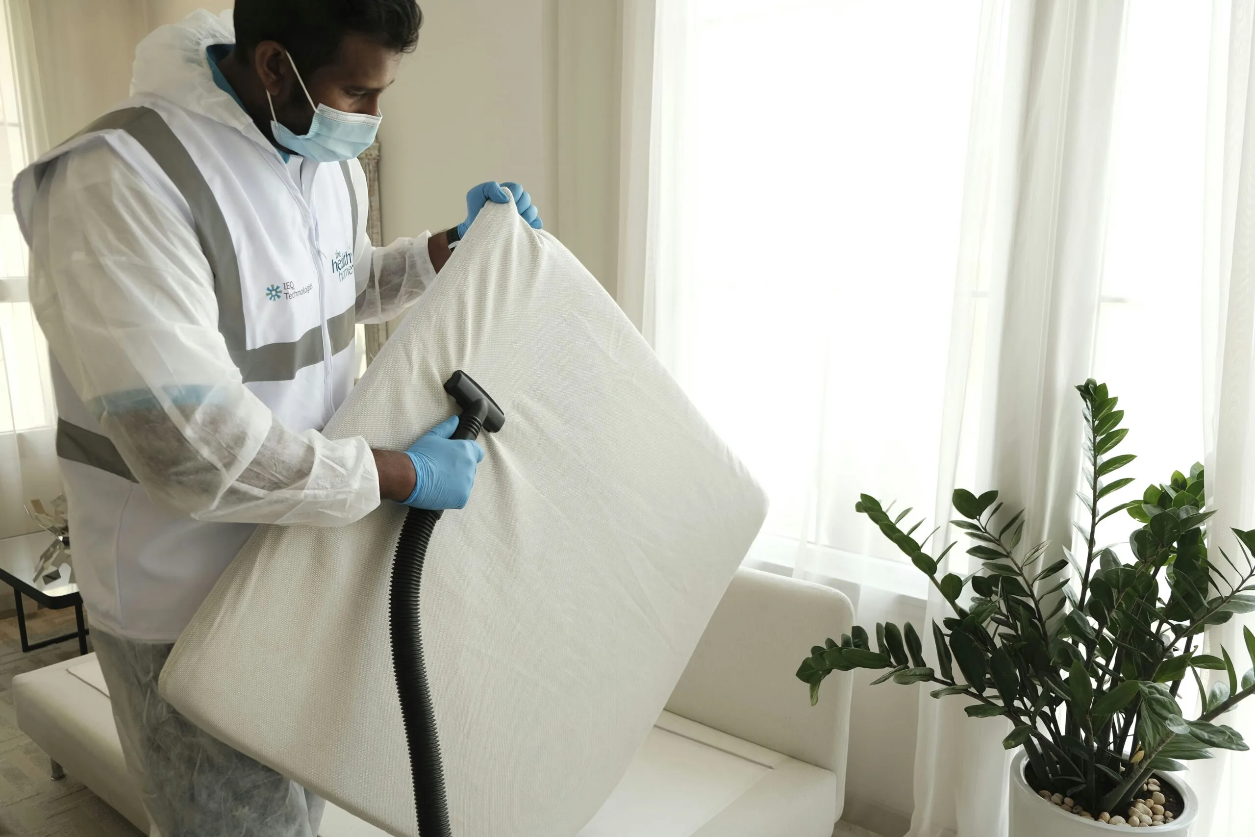 A professional cleaner in a protective suit, mask, and gloves vacuums a sofa cushion in a bright apartment