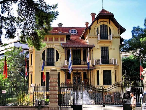 Ornate yellow mansion in Hamidiye with a red-tiled roof, circular windows, and flags behind a wrought-iron fence